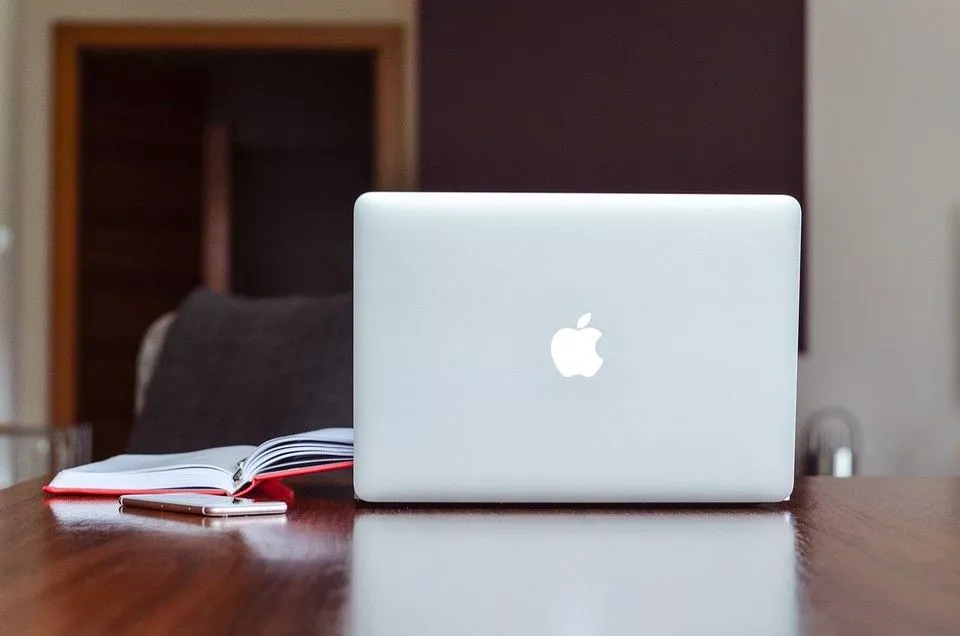 Aufgeklappter Apple-Computer von hinten auf einem Tisch mit Notizbuch und Smartphone daneben.